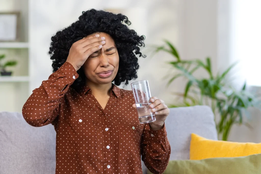 Une femme qui se tient la tête assise dans le canapé qui tient un verre d'eau