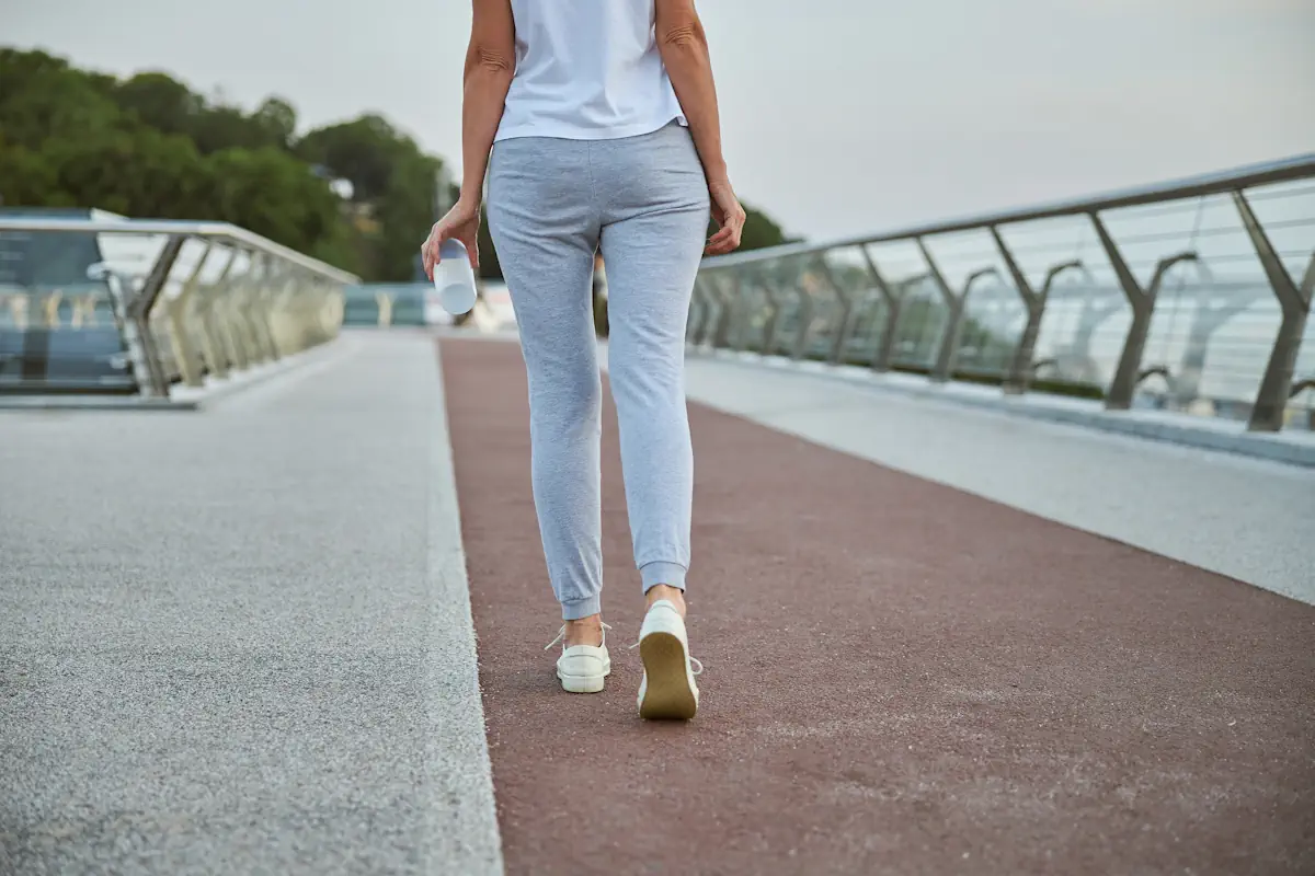 Une femme en pantalon gris et en débardeur blanche qui marche avec une bouteille d'eau à la main